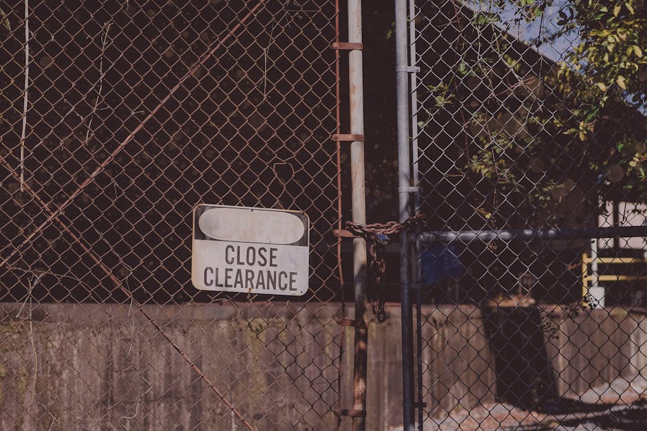 A close-up view of a metal chain-link fence secured with a rusted padlock and chain, blocking access to a dark, densely foliaged area behind it. Attached to the fence is a white rectangular sign with